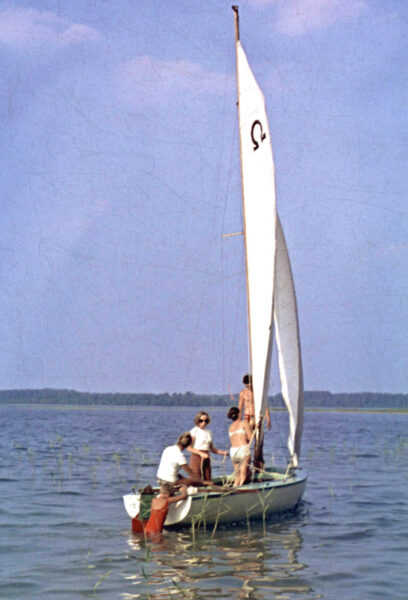 Barndomsfoto av en ung Ariana Ramhage som står i vattnet och skjuter ut en segelbåt från ett grund, medan de övriga passagerarna sitter kvar ombord på båten.