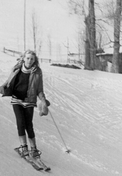 Svartvitt fotografi av en ung Ariana Ramhage som åker skidor i hög fart utför en brant backe, klädd i klassisk skidutrustning med läderpjäxor.