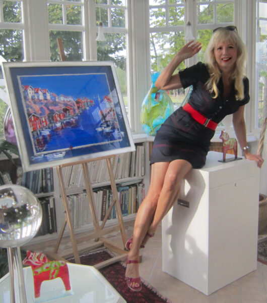 Färgfotografi av konstnären Ariana Ramhage, sittande på en specialdesignad vit trälåda. Hon ler mot kameran. Omkring henne finns inpackad konst och en stor, trälåda för transport till Swedish American Museum i Chicago. I bakgrunden syns målningar i en ateljé.